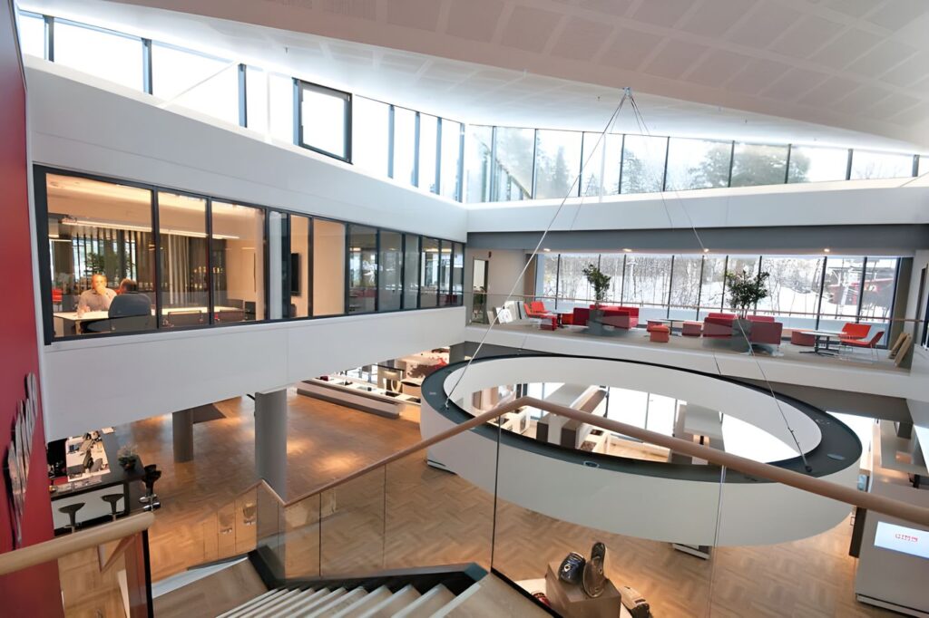 Lyst atrium i kontorbygg med glassvegger, takvinduer og rund åpning mellom etasjene, med møterom og sittegrupper med røde sofaer.
