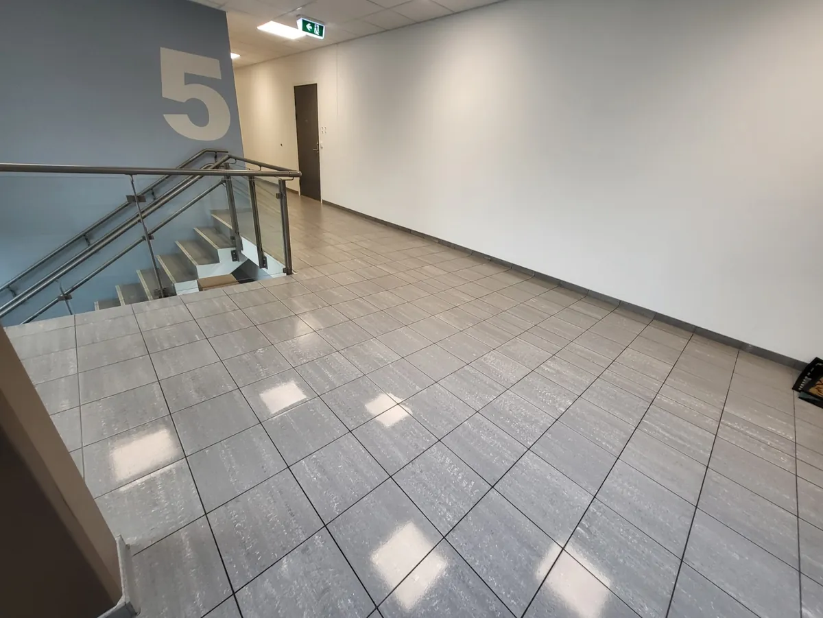 Nyvasket, blankt flisgulv i lys gang ved trappeoppgang i 5. etasje, med glassrekkverk og grå dør i bakgrunnen.