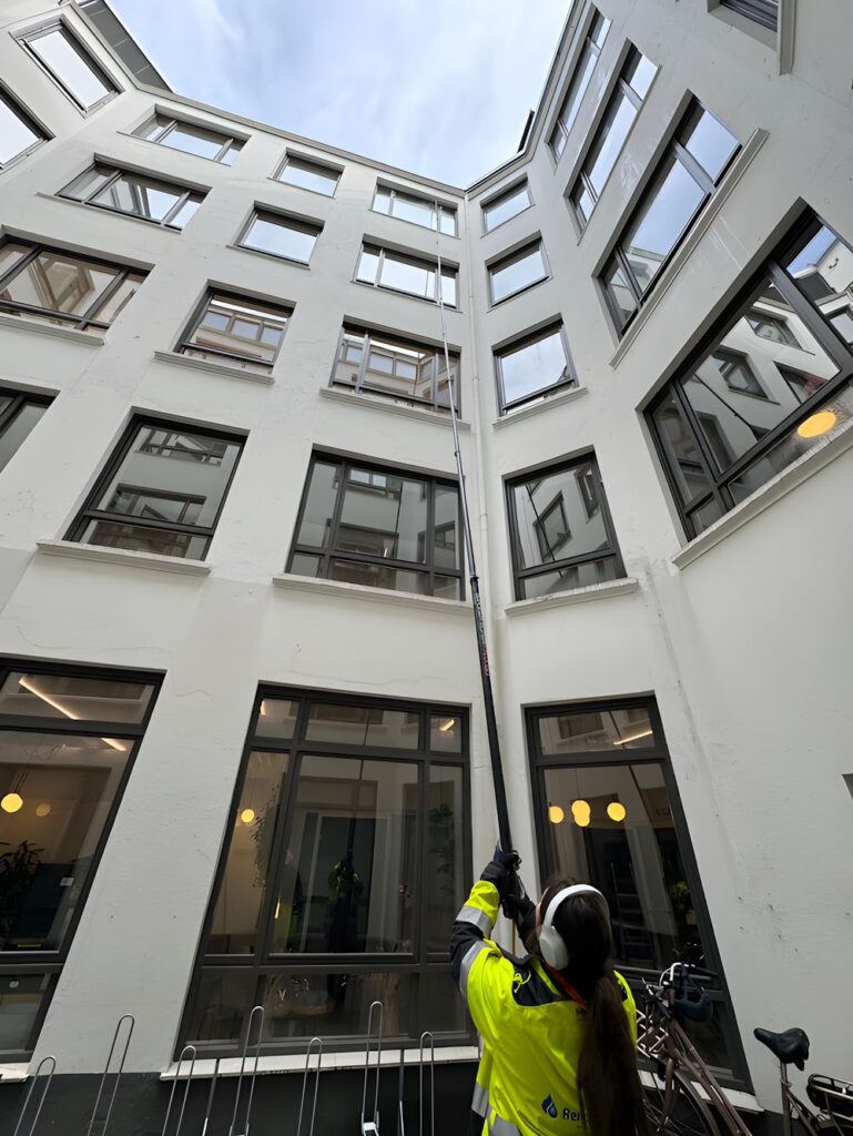 Vindusvasker i gul refleksjakke og hørselvern vasker vinduer høyt oppe med teleskopstang i en hvit bakgård.